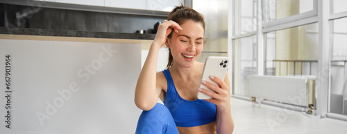 Image of young fit woman wearing sportbra and blue leggings, using smartphone app, does workout from home, sits on floor and follows online training video photo