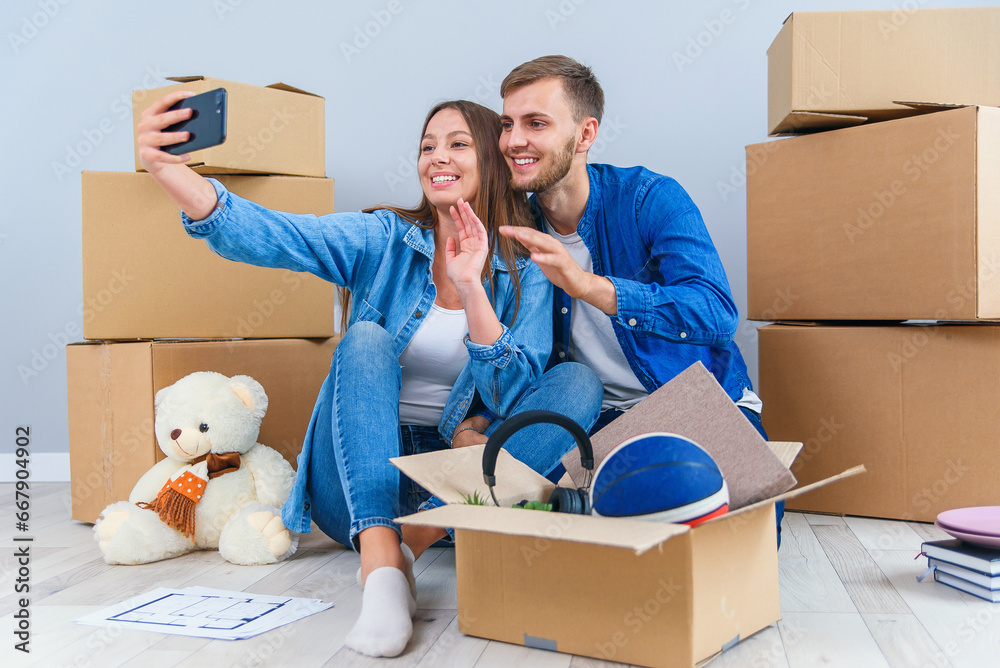 Fototapeta premium Young couple taking selfies in their new home while sitting among cardboard boxes. Happy attractive young couple is moving, making a selfie, cuddling and smiling.