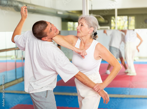 Focused elderly woman performing elbow strike and wristlock, painful control move to immobilize male opponent during self defense training in gym