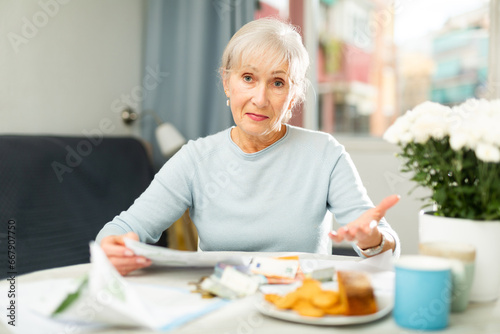 Senior woman sitting at table and looking in camera while counting her budget with bills and documents.