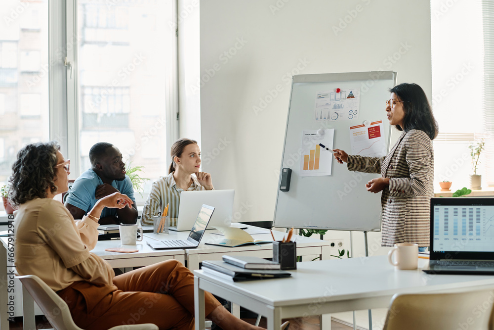 Young confident female analyst pointing at paper document with financial chart on whiteboard while explaining data to group of colleagues Young confident female analyst pointing at paper document with financial chart on whiteboard while explaining data to group of colleagues