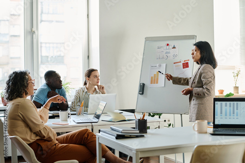 Young confident female analyst pointing at paper document with financial chart on whiteboard while explaining data to group of colleagues