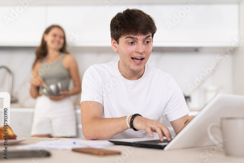 Young smiling woman chatting with young guy working on laptop in kitchen at home