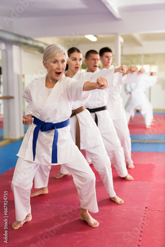 Concentrated aged woman in kimono practicing punches in gym during group martial arts workout. Shadow fight, combat sports training concept