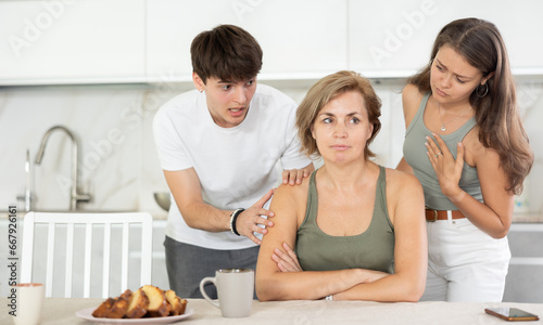 Middle-aged woman sits sad and thoughtful near kitchen table, adult children, guy and girl ask relative about problems and offer help.