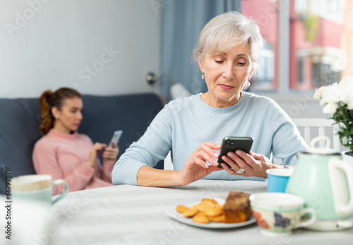 Senior woman sitting at table at home and using her smartphone. Her granddaughter sitting on sofa in background and using smartphone too.