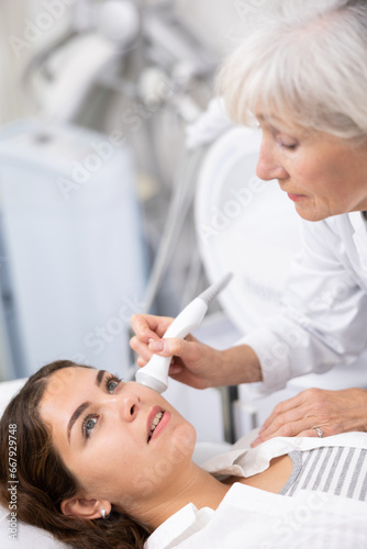 Pleased young woman lying on clinical chair in aesthetic cabinet during face laser therapy procedure