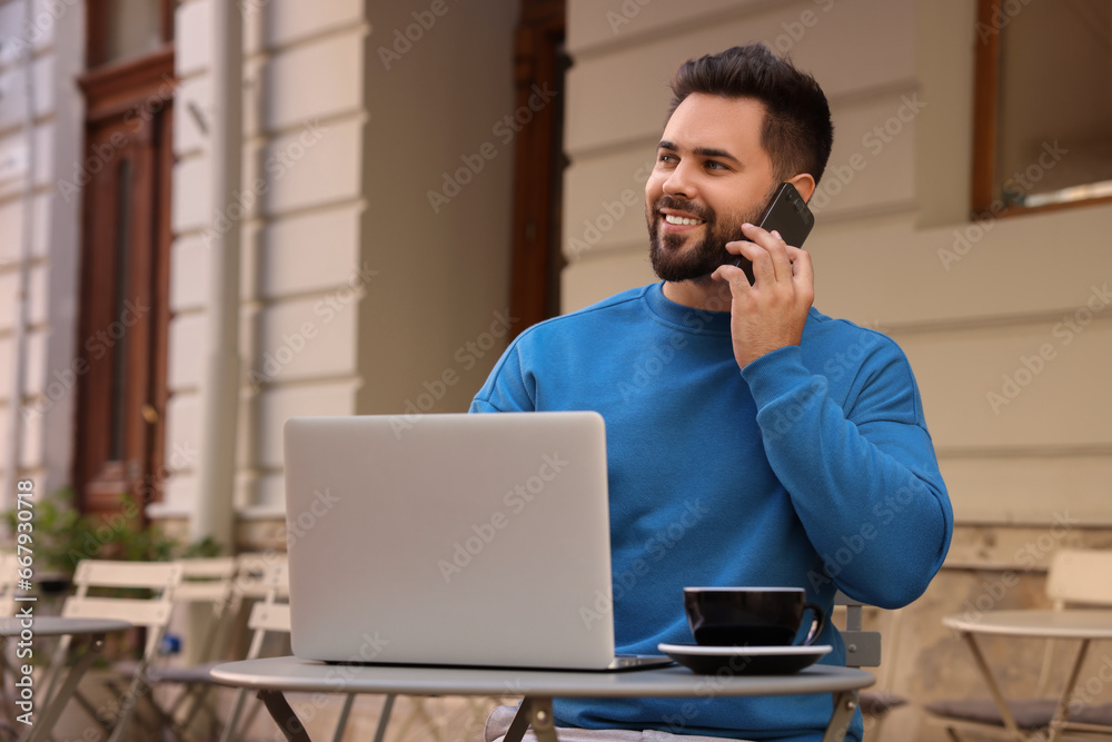 Handsome young man talking on smartphone while using laptop at table in outdoor cafe Handsome young man talking on smartphone while using laptop at table in outdoor cafe