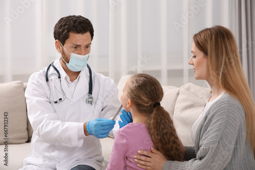 Doctor in medical mask examining girl`s oral cavity with tongue depressor near her mother indoors