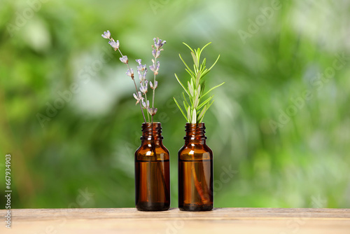 Bottles with essential oils, lavender and rosemary on wooden table against blurred green background