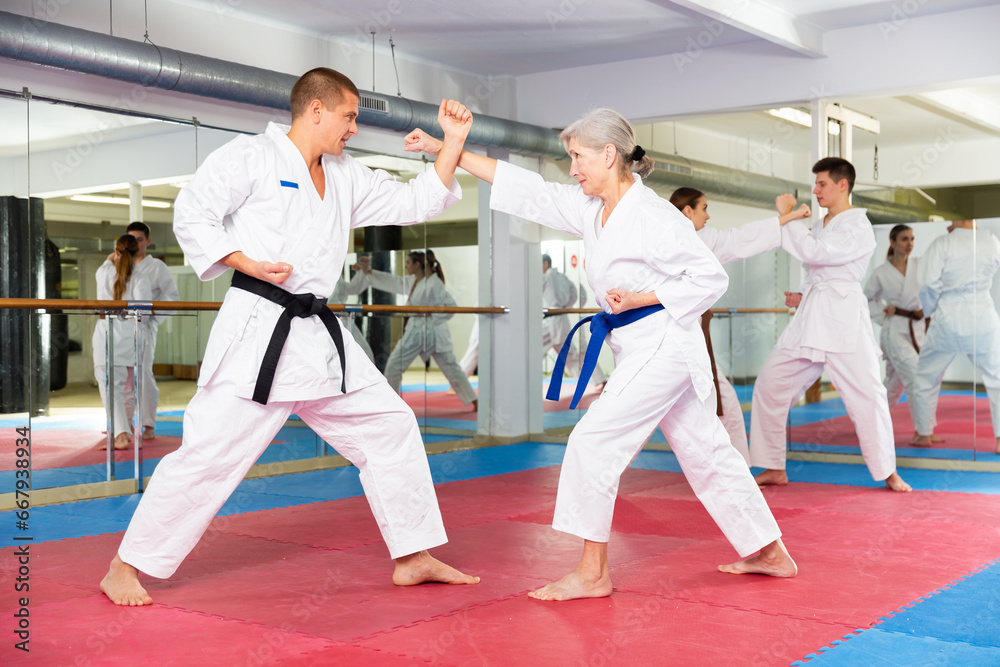 Caucasian man and senior woman in kimono sparring during group karate training. Woman and teenage boy fighting in background.