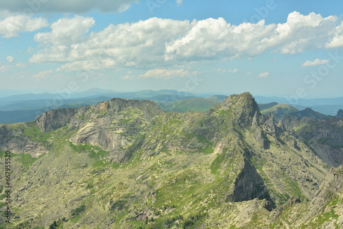A high rocky massif with peaked peaks partially overgrown with grass under a cloudy summer sky.