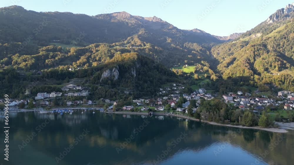 An aerial view above Walensee captures a small marina and residential houses with the majestic green-dressed mountain Alps reflected beautifully in the water on a sunny day An aerial view above Walensee captures a small marina and residential houses with the majestic green-dressed mountain Alps reflected beautifully in the water on a sunny day