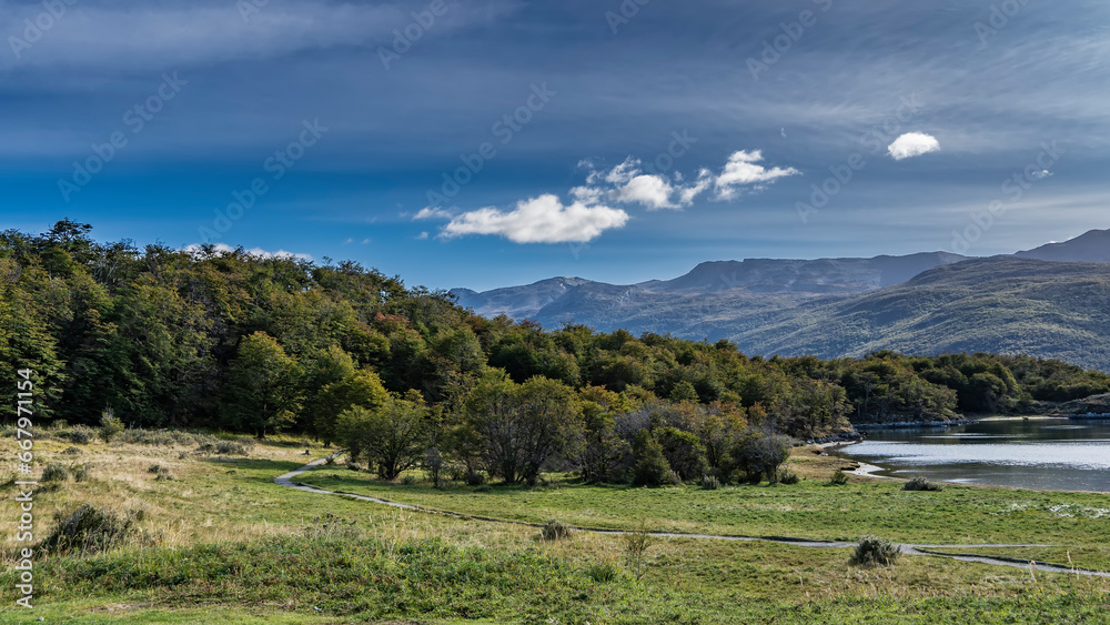 Beautiful landscape of the nature reserve. The path winds through a green meadow next to the lake and goes into the forest. Picturesque mountains against a blue sky, clouds. Argentina. Lapataia Bay.