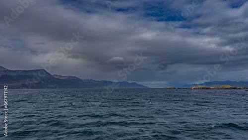 The wide blue Beagle Channel stretches to the horizon. Small waves on the water. Islands in the distance. A picturesque mountain range against a blue sky and clouds. Argentina. Tierra del Fuego 