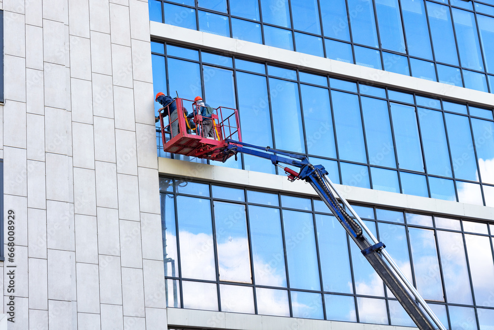 External glass facade window washers, while in a construction lifting cradle, clean the glass facade of a building on a clear day.