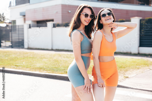 Two young beautiful smiling female in trendy summer cycling shorts, top clothes. Sexy carefree women posing in street at sunny day. Positive models having fun. Cheerful and happy. In sunglasses