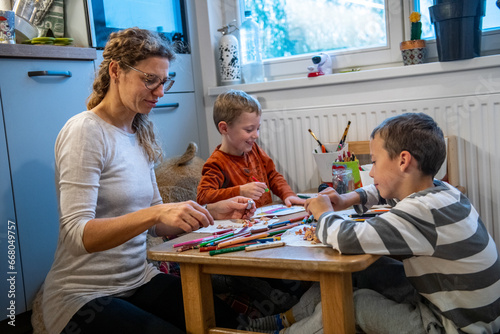 A mother and two children are drawing with crayons. they are sitting at the table. They are getting ready for school. They are sitting behind floor sitting desk