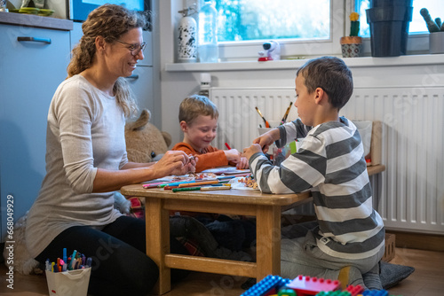 A mother and two children are drawing with crayons. they are sitting at the table. They are getting ready for school. They are sitting behind floor sitting desk