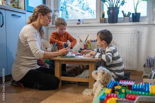 A mother and two children are drawing with crayons. they are sitting at the table. They are getting ready for school. They are sitting behind floor sitting desk
