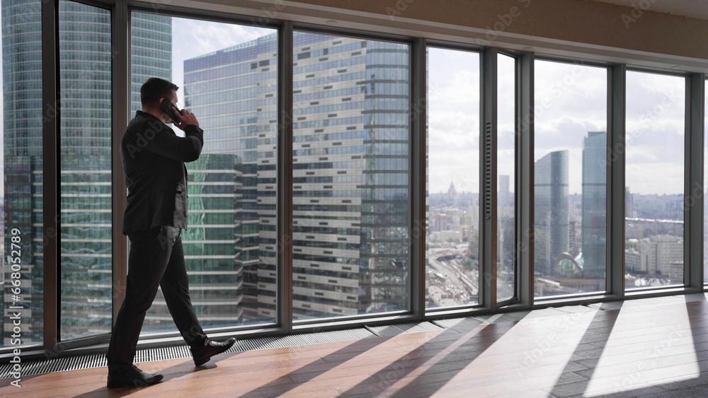 A young businessman uses a mobile phone in the corridor of a skyscraper. A businessman is talking on the phone in the corridor, skyscrapers outside the window. A young businessman uses a mobile phone in the corridor of a skyscraper. A businessman is talking on the phone in the corridor, skyscrapers outside the window.
