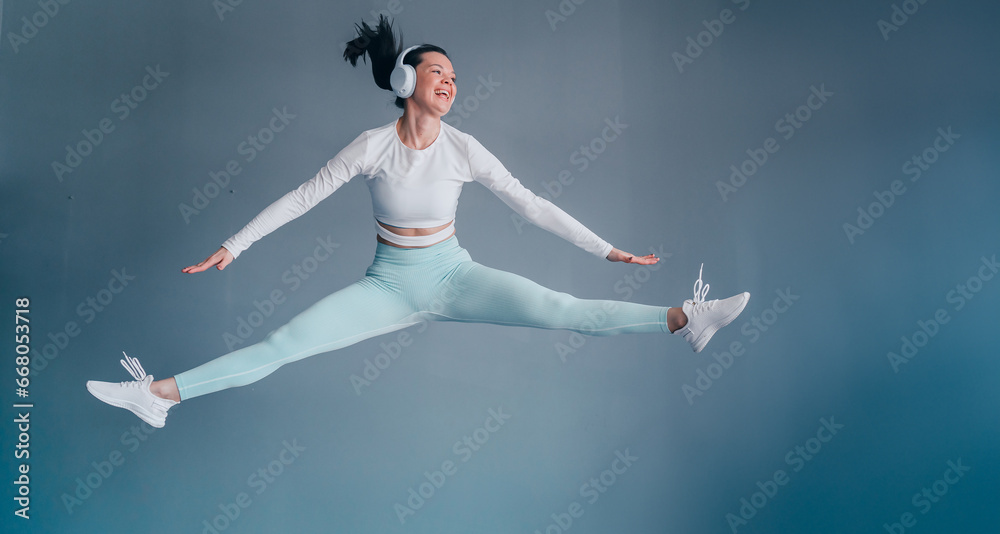 Excited young woman in sportswear jumping high at gym listening motivating music using headphones looks away toothy smiling. Sport club. Excited young woman in sportswear jumping high at gym listening motivating music using headphones looks away toothy smiling. Sport club.