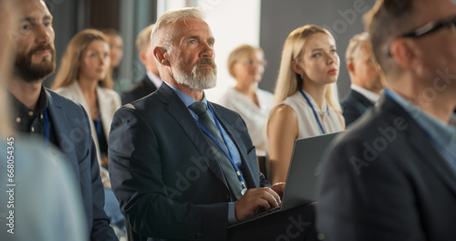 Senior Male Investor Sitting In Crowd And Using Laptop Computer At International Technology Conference. Caucasian Man Listening To Keynote Presentation About New Innovative Startup Company Service.