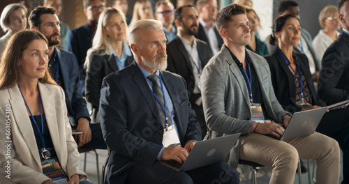 Senior Caucasian Man Sitting in a Crowded Audience at a Business Forum. Corporate Delegate Using Laptop Computer. Successful Male CEO Attending Business Conference, Listening To Presentation.