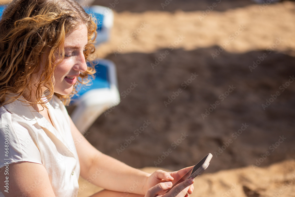 A woman smiling while using a smartphone on the beach.