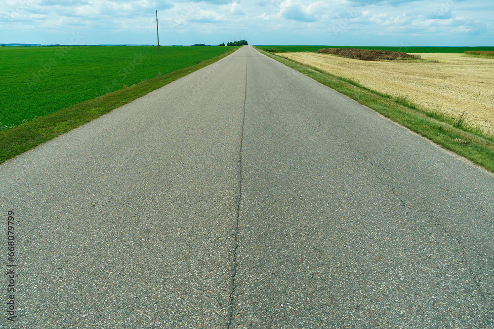 An empty roadway outside the city passes by agricultural fields. A highway with a new asphalt surface on the background of fluffy clouds. Paved road on a sunny day without cars. An empty roadway outside the city passes by agricultural fields. A highway with a new asphalt surface on the background of fluffy clouds. Paved road on a sunny day without cars.