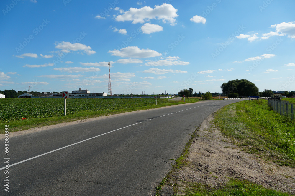 An empty roadway outside the city passes by agricultural fields. A highway with a new asphalt surface on the background of fluffy clouds. Paved road on a sunny day without cars.