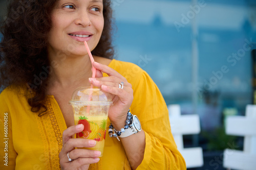 Close-up young adult multi-ethnic woman drinking fresh juice outdoor, pensively looking aside a copy advertising space