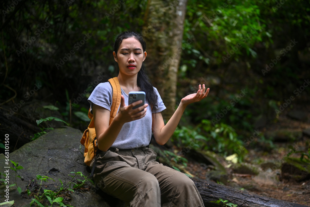 Upset female hiker with backpack sitting on rocks looking for connection to the internet on smartphone