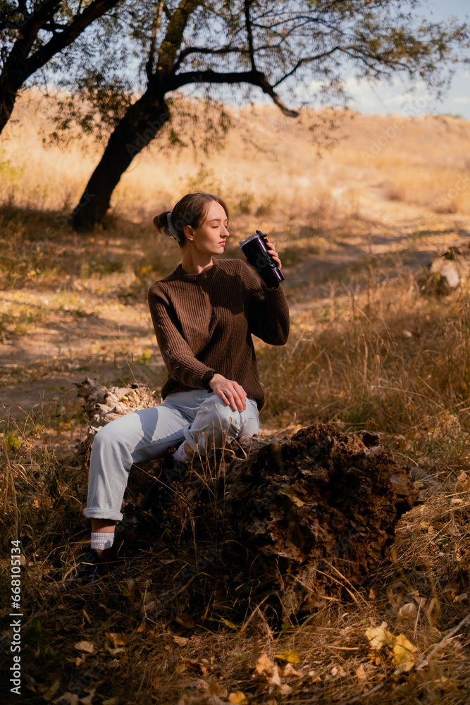 young woman relaxing outdoors on an autumn day, traveling to the forest, sitting on a wooden log in the forest and drinking hot tea, unity with nature, searching for the meaning of life