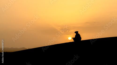 Silhouette of unidentified Berber man sitting, watching sunset on sand dunes at beautiful sunset in Sahara Desert, Morocco