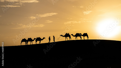 Silhouette of two Berber men leading camel caravans come across each other on sand dunes during sunset in Sahara Desert  Morocco