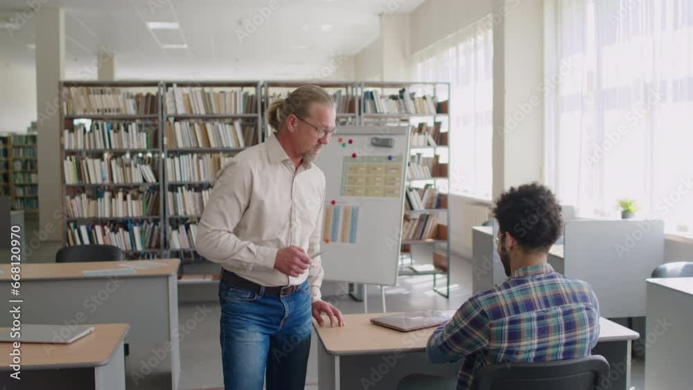Medium shot of mature Caucasian male English teacher checking and helping multiethnic group of migrant students sitting at single desks in library and studying language Medium shot of mature Caucasian male English teacher checking and helping multiethnic group of migrant students sitting at single desks in library and studying language