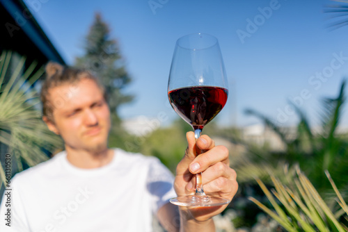 Tableau sur toile A young charming man holds out a glass of wine to the camera and carefully exami