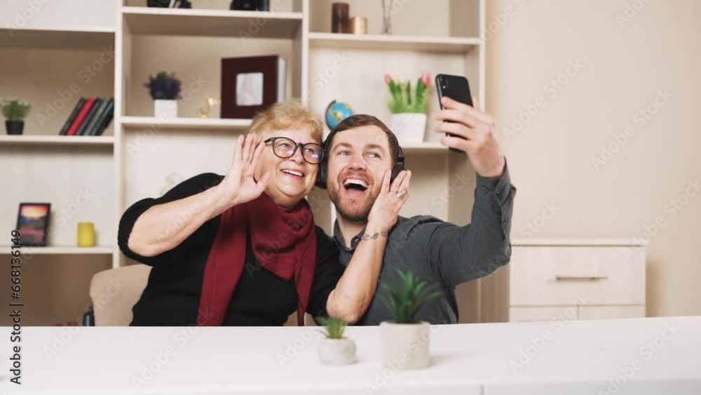 young man in wireless earbuds takes a selfie on a smartphone with an older woman wearing glasses at home young man in wireless earbuds takes a selfie on a smartphone with an older woman wearing glasses at home
