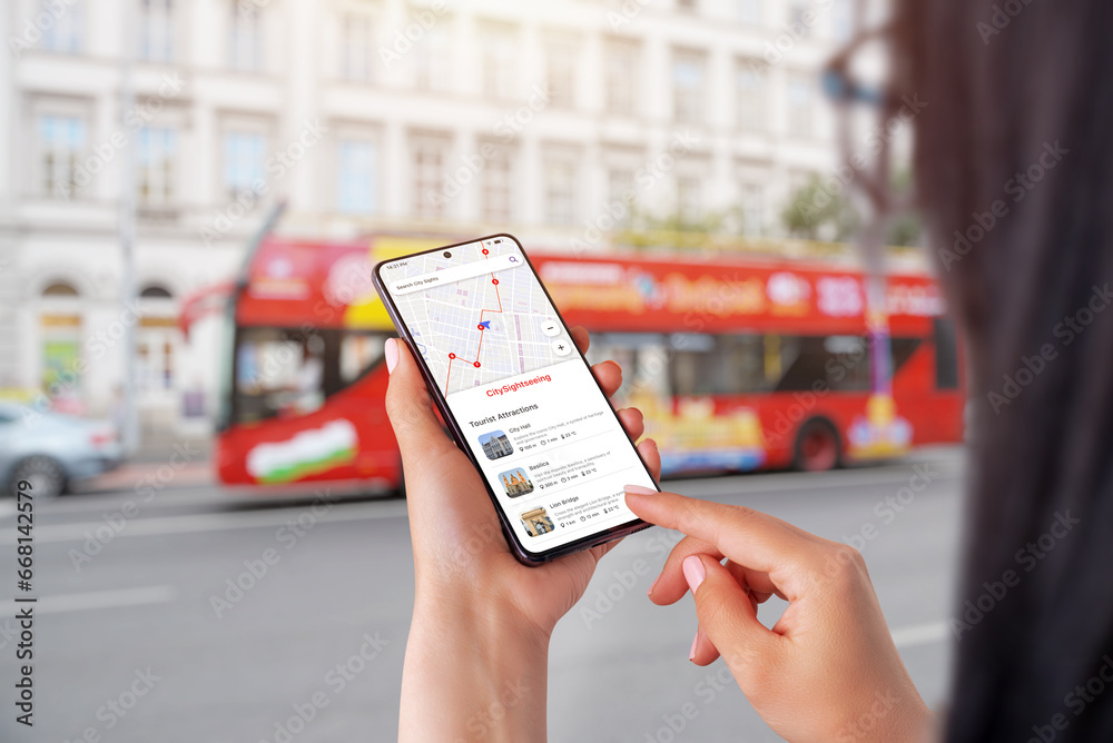 City Sightseeing app on smartphone in woman hands. City tourist bus in background - obrazy, fototapety, plakaty 