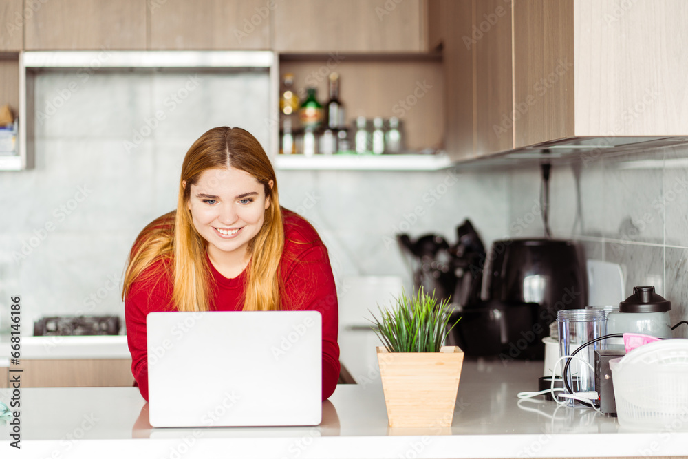 Beautiful, smiling woman standing, using laptop, working in kitchen in modern apartment, at home Beautiful, smiling woman standing, using laptop, working in kitchen in modern apartment, at home