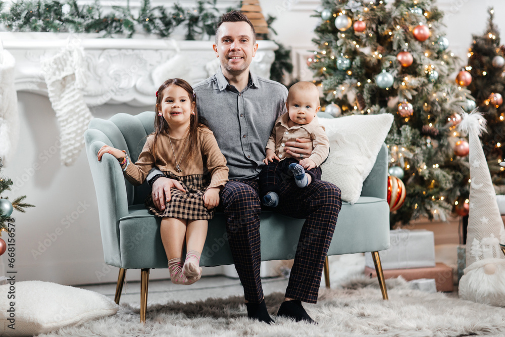 Dad spends time with his little children in front of a Christmas tree and Christmas decorations. Happy father and his son and daughter play together. Dad hugs his happy children Dad spends time with his little children in front of a Christmas tree and Christmas decorations. Happy father and his son and daughter play together. Dad hugs his happy children