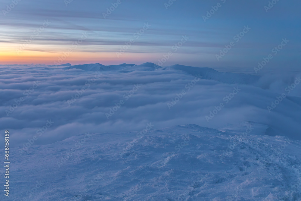 Winter landscape in the mountains at sunset of the day. Clouds flowing over the ridge. Mountains and sunset can be seen in the distance.