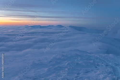 Winter landscape in the mountains at sunset of the day. Clouds flowing over the ridge. Mountains and sunset can be seen in the distance.