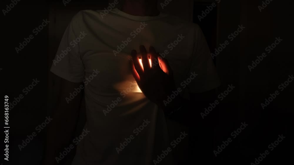 A man in a white Tshirt in a dark room holds his hand on his heart
