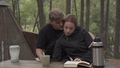 Glamping wooden teepee, the bride and groom in autumn clothes is sitting outdoors on a wooden table while relaxing reading a book and drinking coffee.