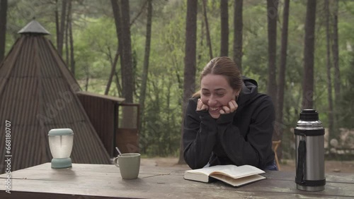 Glamping wooden teepee, the bride and groom in autumn clothes is sitting outdoors on a wooden table while relaxing reading a book and drinking coffee.