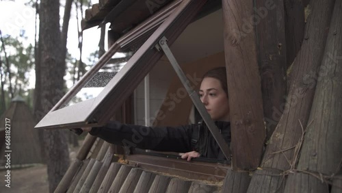 Girl in hat and autumn clothes opens the window of a log cabin during autumn.