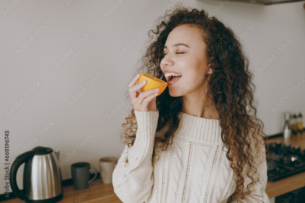 Close up young vegetarian housewife woman of African American ethnicity wear casual clothes sweater eat bite half of orange stand near table in light kitchen at home. Lifestyle cooking food concept. Close up young vegetarian housewife woman of African American ethnicity wear casual clothes sweater eat bite half of orange stand near table in light kitchen at home. Lifestyle cooking food concept.