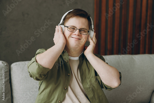 Young man with down syndrome wear glasses casual clothes listen to music in headphones sits on grey sofa couch stay at home flat rest spend free time in living room. Genetic disease world day concept. photo